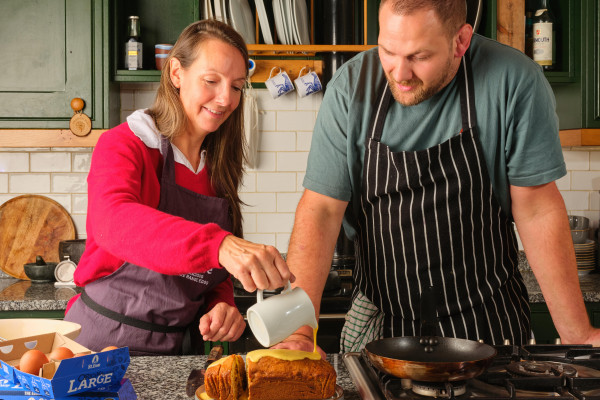 A woman and a man stood behind a counter in a kitchen, pouring custard over a cake